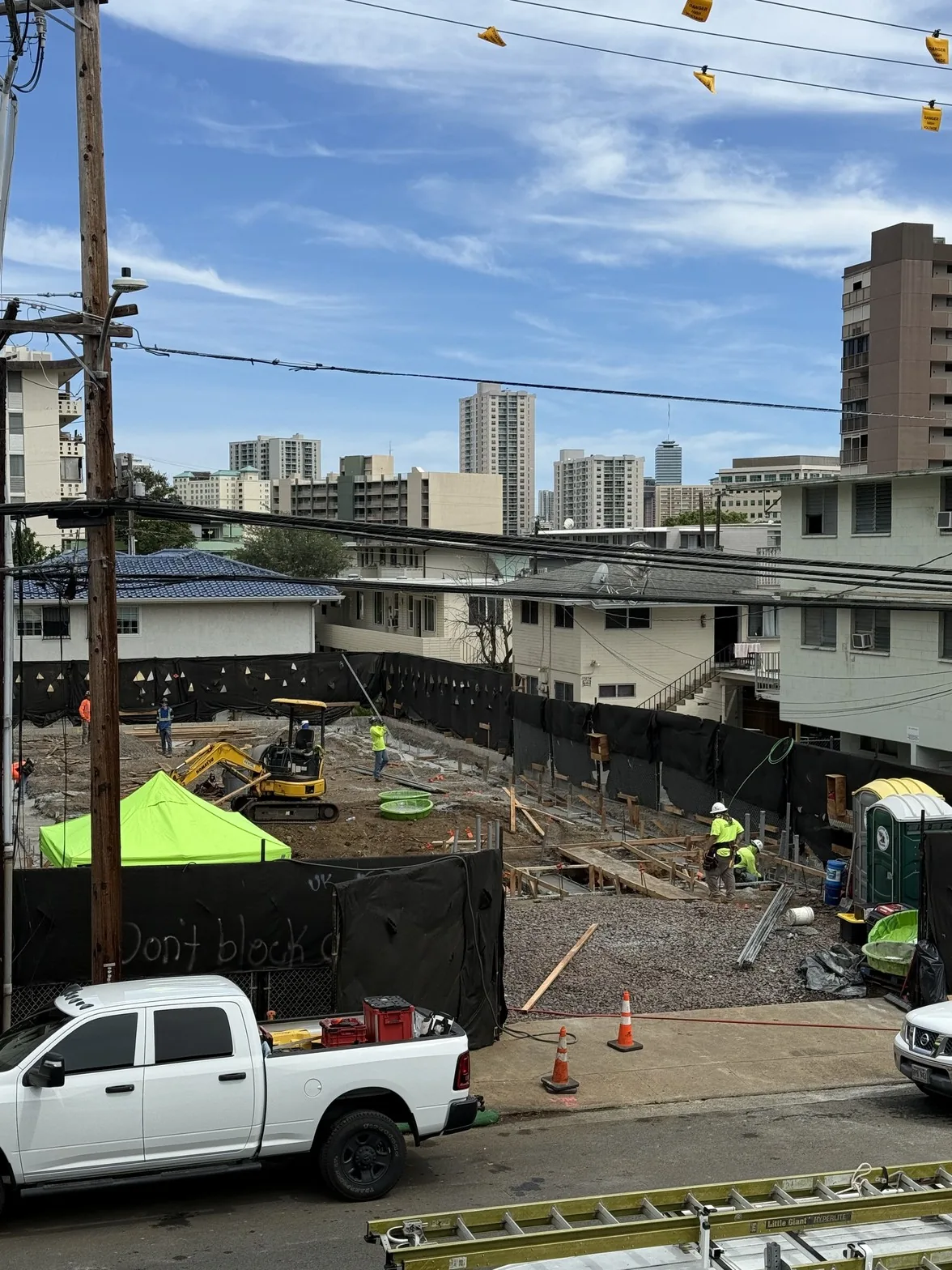 ホノルル都市部の既成市街地に建つ建設現場の全景と、周囲の既存住宅およびダウンタウンのスカイライン
